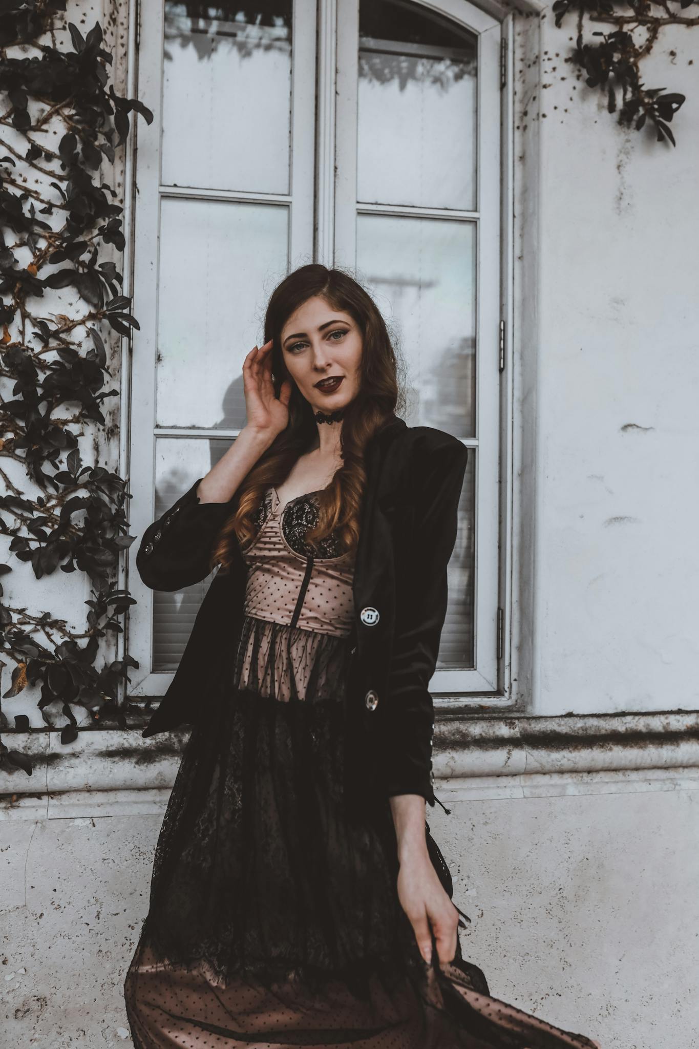 Fashionable woman in black dress poses by a vintage window, exuding elegance.
