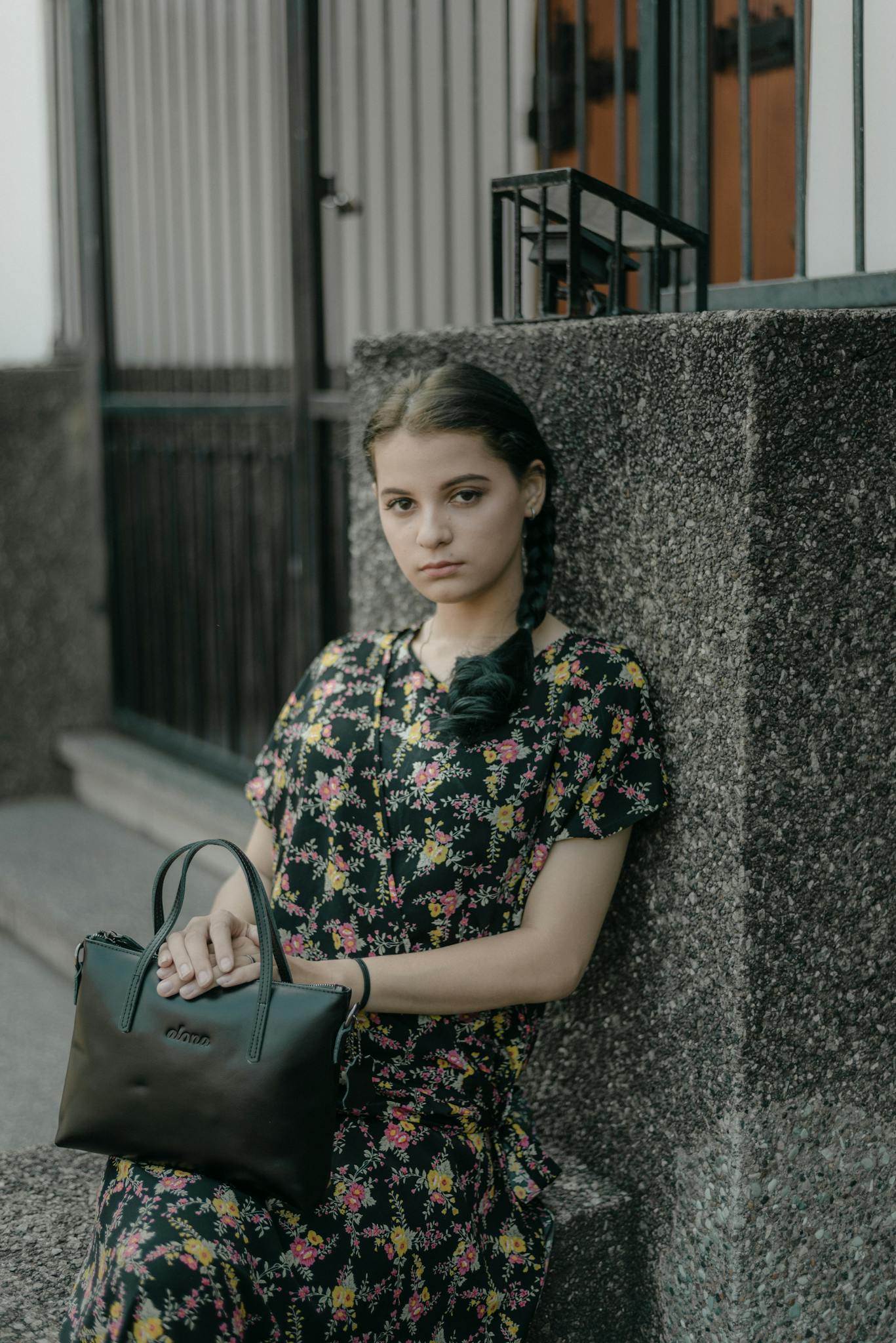 Woman in floral dress and braided hair sitting on a concrete bench with a black bag.