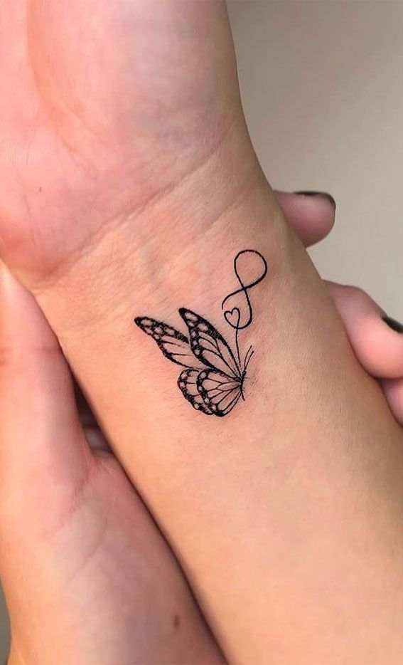 Cute arm tattoos for women featuring a delicate, small black-line butterfly with open, patterned wings on the wrist. The butterfly's flight path continues into a fine-line design incorporating a small heart and an infinity symbol, representing eternal love and transformation. This feminine and trending style is perfect for a wrist placement.