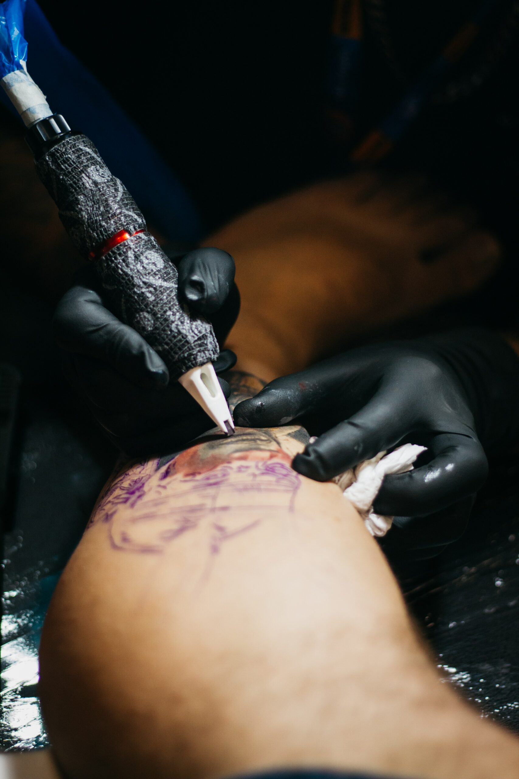 Text Close-up of a tattoo artist in black gloves using a tattoo machine on a person's arm, with a purple stencil visible, illustrating the start of a new design for a cute arm tattoo for women.